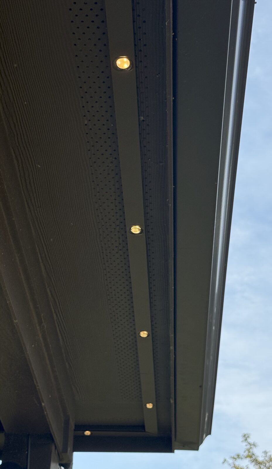 Black color-matched permanent lighting track with warm-white pucks mounted on a dark soffit, seen during the day