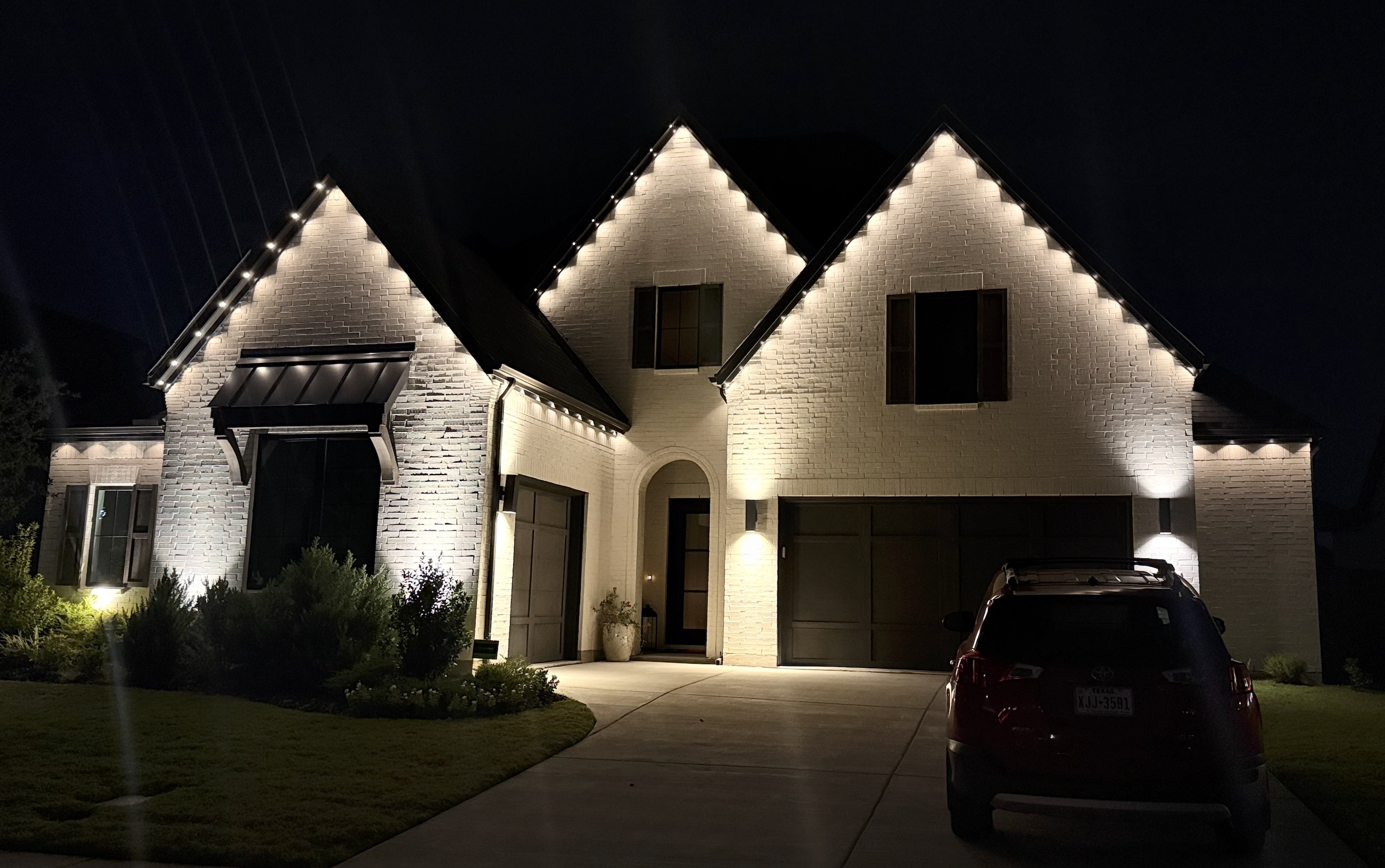 White brick home with three gables outlined in warm white permanent eave lighting at night in Texas